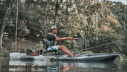 Man casting a fishing pole while sitting in a lake on his grey fishing kayak with trees in the background.