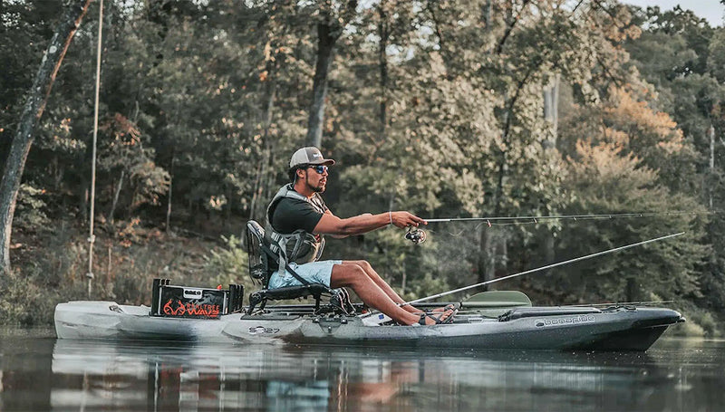 Man casting a fishing pole while sitting in a lake on his grey fishing kayak with trees in the background.