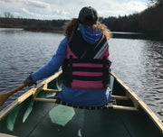 Woman wearing a pink life jacket paddling her green canoe down a calm river.