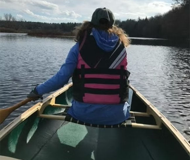 Woman wearing a pink life jacket paddling her green canoe down a calm river.