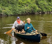 A man and a woman with a dog paddling their black canoe down a calm river.