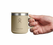 Hand holding up a small tan travel mug with a Hydroflask logo on it on a white background.