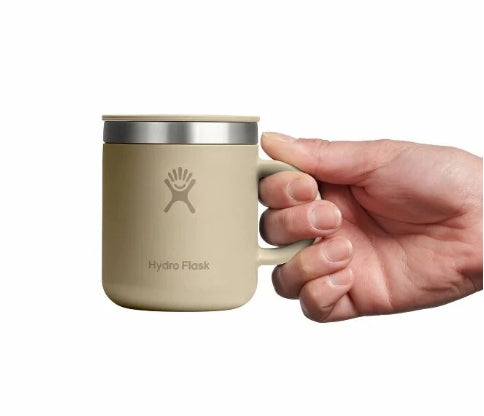 Hand holding up a small tan travel mug with a Hydroflask logo on it on a white background.