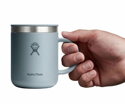 Hand holding up a small light grey travel mug with a Hydroflask logo on it on a white background.
