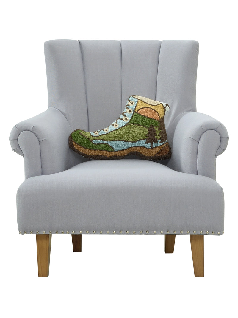 Boot shaped pillow with a forest pattern on it resting on a gray chair against a white background. 