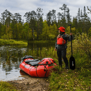Woman standing on a riverbank next to her red inflatable kayak with trees in the background.