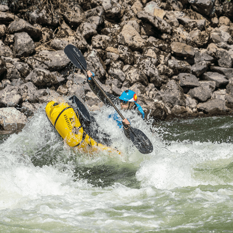 Man in a yellow inflatable kayak paddling through rough waters with rocks in the background.