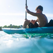 Man paddling a blue and white inflatable kayak in a lake with the sun in the background. 