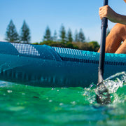 Side view of a person in a blue inflatable kayak with white accents paddling down a river. 