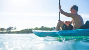 Young man rowing a blue and white inflatable kayak on a very sunny day with trees in the background. 