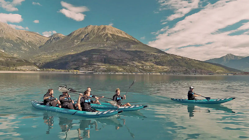 Three blue and white kayaks filled with happy people rowing down a crystal clear river with mountains in the background.