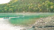 Three blue and white inflatable kayaks filled with happy people rowing down a crystal clear river with mountains and forest in the background. 