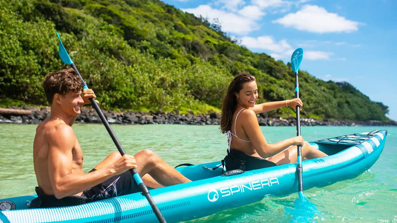 A man and a woman in a blue and white colored inflatable kayak rowing with blue and black paddles down a river.