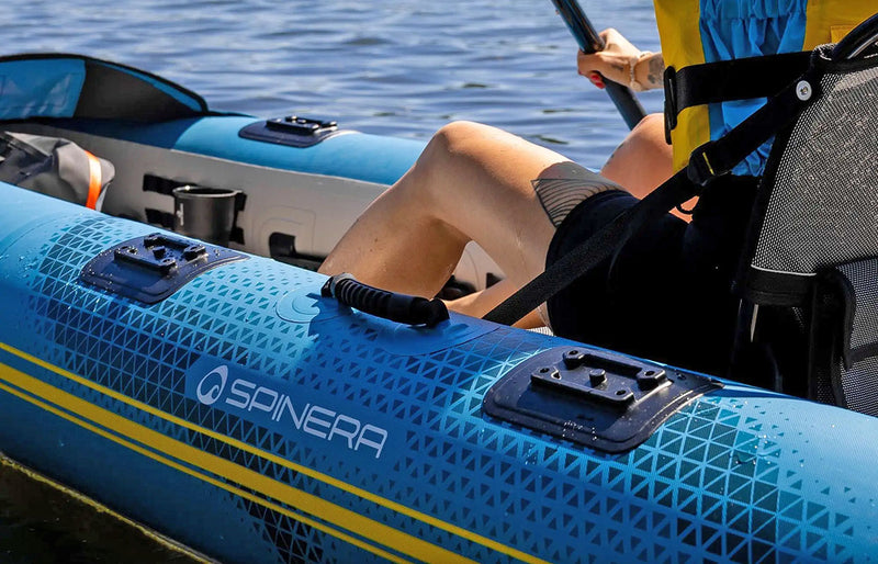 Close up of a blue inflatable kayak with yellow accents and a Spinera logo on the side in a lake. 