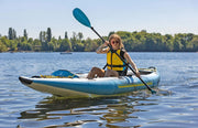 Woman with cool sunglasses in a blue inflatable kayak with yellow accents paddling in a lake. 