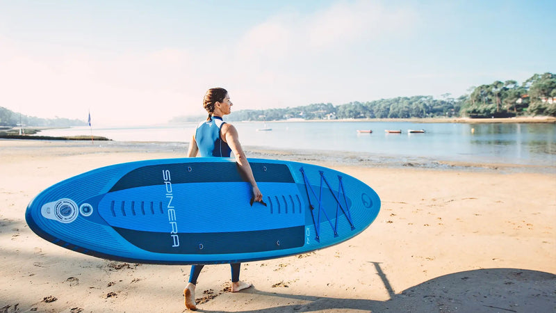 Person holding a blue inflatable stand-up paddleboard on a sandy beach with water and trees in the background.