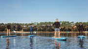People paddleboarding on a calm body of water with trees and clear sky in the background.