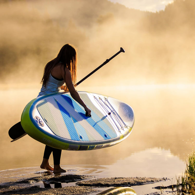 Woman carrying her blue and yellow paddleboard along the shore of a lake. 