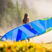 Woman carrying a blue paddleboard with yellow accents through a field. 