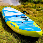 Close up of a blue paddleboard with yellow accents in some grass near a lake. 
