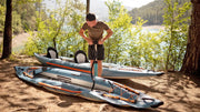 Man pumping air into a blue inflatable kayak with orange and white accents in a clearing by a lake. 