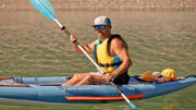 Man in sunglasses rowing down a river in a blue inflatable kayak with orange and white accents. 