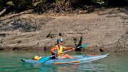 Man with hat and sunglasses paddling down a river in a blue inflatable kayak with orange and white accents. 