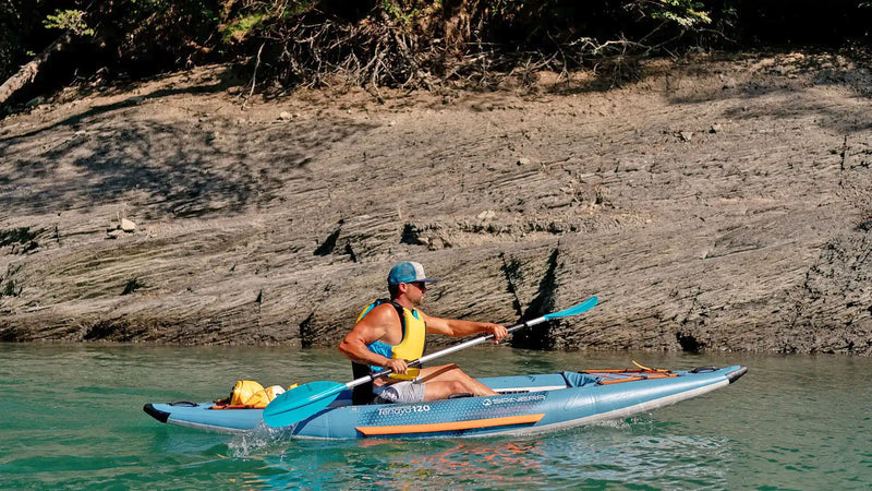 Man with hat and sunglasses paddling down a river in a blue inflatable kayak with orange and white accents. 