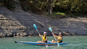 A man and a woman rowing down a river in a blue and orange tandem inflatable kayak. 