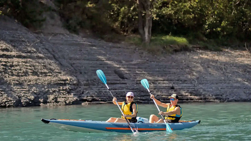 A man and a woman rowing down a river in a blue and orange tandem inflatable kayak. 