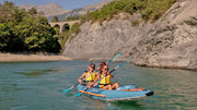 Two smiling women in a blue kayak with orange accents rowing down a river with mountains and a bridge in the background. 