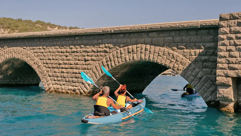 A man and a woman in a blue kayak with orange accents rowing through a tunnel under a bridge. 