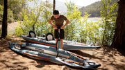 Man using a hand pump to put air in a blue inflatable kayak with orange accents in a clearing next to a lake. 