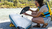 Woman installing replaceable fin onto a blue and white inflatable kayak next to a river. 