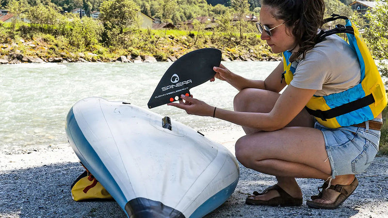 Woman installing replaceable fin onto a blue and white inflatable kayak next to a river. 