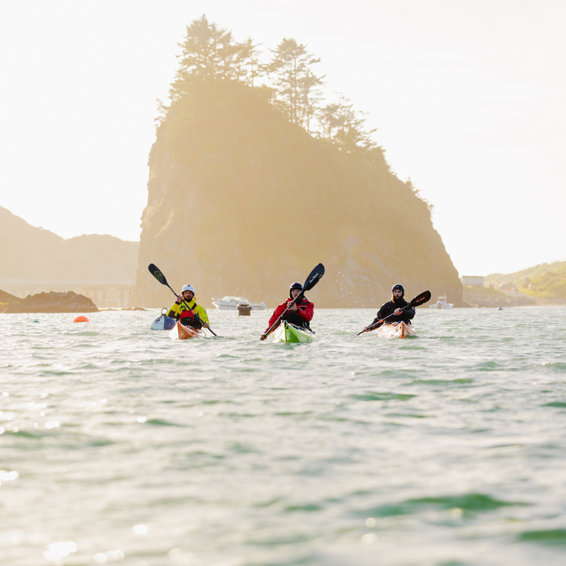 Three kayakers paddling on a calm body of water with a scenic background.