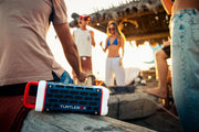 Turtlebox speaker on a wooden surface with people in the background at sunset.