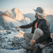 Man with fun hat sitting at the edge of a mountain cliff with a bag of freeze dried blueberry peach crisp at his side.
