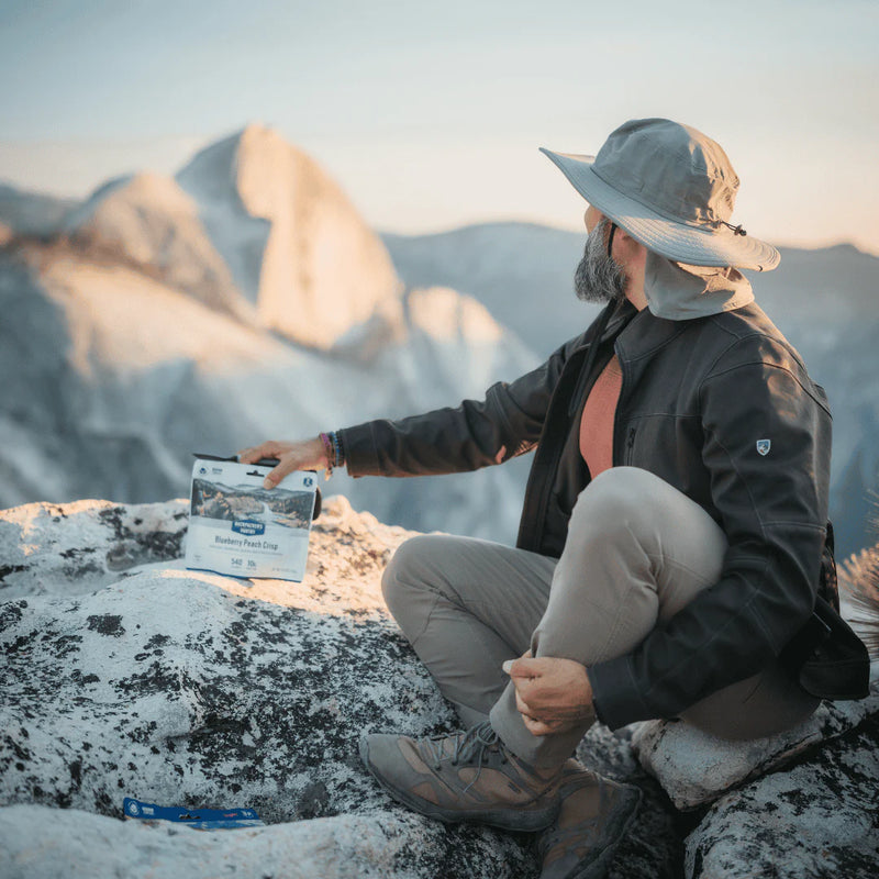 Man with fun hat sitting at the edge of a mountain cliff with a bag of freeze dried blueberry peach crisp at his side.