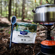 Bag of blueberry peach crisp oatmeal sitting on the ground in a forest next to a blue camp spoon and a camp stove. 