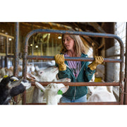 A woman wearing a teal zip-up jacket and tan work gloves stands behind a metal gate in a barn, holding goats inside a pen.