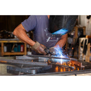 A welder in a workshop wearing protective gloves and a welding helmet, actively welding metal pieces with bright blue light and orange sparks flying. The welder is wearing a dark blue t-shirt.
