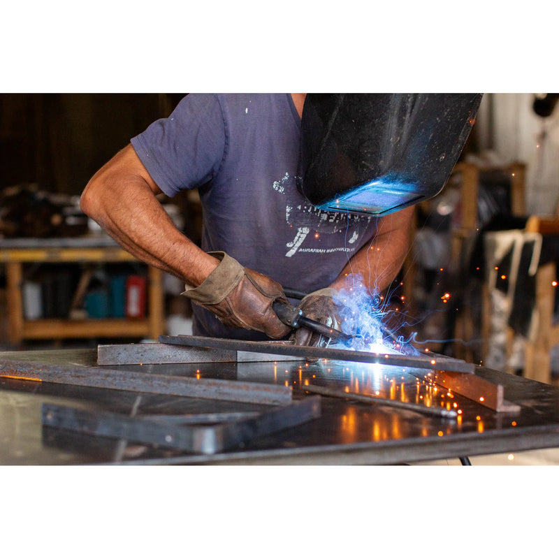 A welder in a workshop wearing protective gloves and a welding helmet, actively welding metal pieces with bright blue light and orange sparks flying. The welder is wearing a dark blue t-shirt.