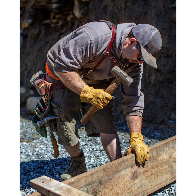 A man wearing a gray hoodie, brown shorts, and yellow leather work gloves uses a sledgehammer to lift a wooden beam in an outdoor construction setting.