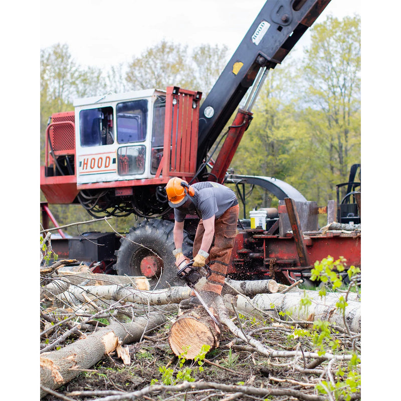 A worker wearing safety gear uses a chainsaw to cut logs in a logging operation, with a large red and black logging machine and trees in the background.