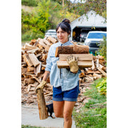 A woman outdoors carrying stacked firewood on her back and holding a single log in one hand, wearing a light blue denim shirt, dark blue shorts, and tan work gloves. Piles of chopped wood and a white shed are visible in the background.
