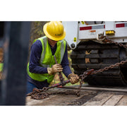 A construction worker wearing a yellow hard hat, high-visibility safety vest, and work gloves is operating a chain tensioner on a flatbed truck next to a white tracked vehicle.
