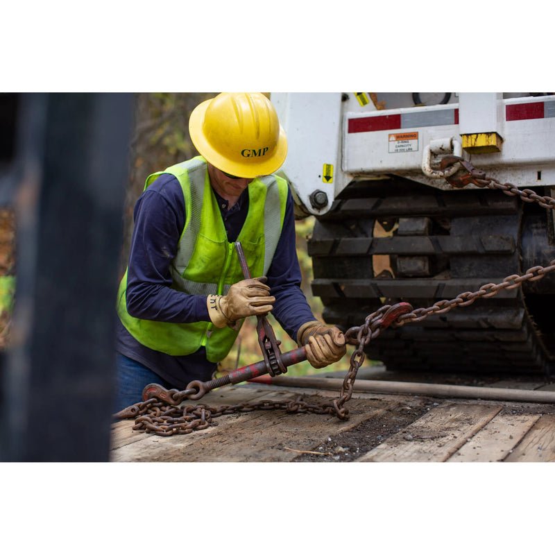 A construction worker wearing a yellow hard hat, high-visibility safety vest, and work gloves is operating a chain tensioner on a flatbed truck next to a white tracked vehicle.