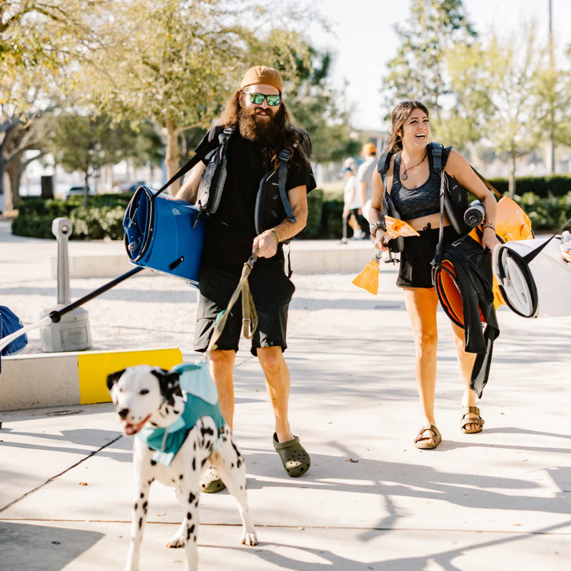 Two people walking a dog on a leash while carrying Tucktec kayaks folded for transport from a shoulder strap, in an urban setting.