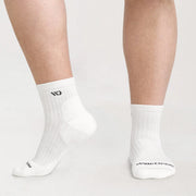 White crew socks worn on a person's feet, shown against a clean light gray studio background. The socks have black printed branding near the toe and ankle.